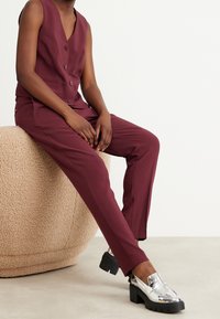 Burgundy ärmlös väst med knappar, parat med matchande skräddarsydda byxor. Skorna har metallisk silverfärgade loafers med kraftiga sulor.