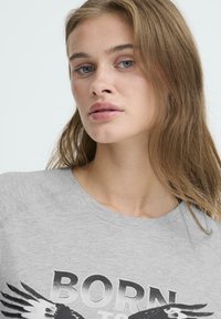 Camiseta de algodón gris de manga corta, con un atrevido estampado gráfico en blanco y negro que dice "NACIDO para..." y un diseño de alas de águila.