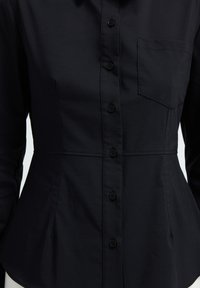 Camicia nera con bottoni, con vita aderente, colletto e una tasca sul petto. Presenta una texture liscia e sei bottoni frontali.