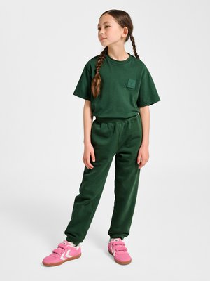 Ung pige med fletninger iført mørkegrøn t-shirt og bukser med pink sportssko, står med afslappede hænder mod hvid baggrund.