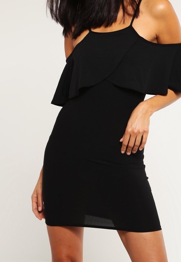 Femme portant une robe mini noire sans manches avec un décolleté asymétrique volanté décolleté épaules sur un fond uni.