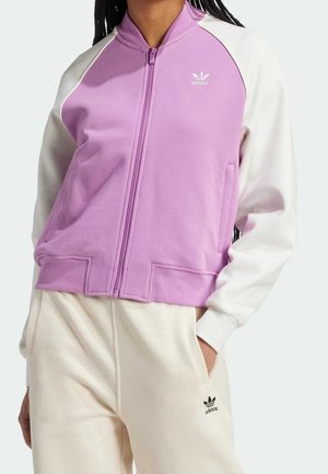 Persoon draagt een paars-witte Adidas jas met rits en witte Adidas trainingsbroek, hand in de zak, bij de borst en heupen bijgesneden.