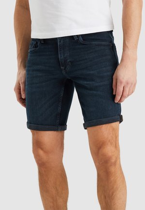 Un homme portant un short en denim bleu foncé retroussé et un t-shirt blanc sur un fond clair uni.