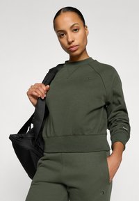 Zöld pulóver, kötött anyagból készült, kerek nyakkivágással, bordázott mandzsettákkal és logó részlettel. A modellen fekete táska van.