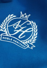 Modrá tkanina s bielym logom, ktoré obsahuje korunu, vavrínové listy a text "NOBLE HERITAGE" na textúrovanej ploche.