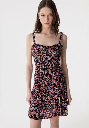 Giovane donna con capelli castani lunghi che indossa un abito nero senza maniche con stampa floreale multicolore su sfondo semplice.