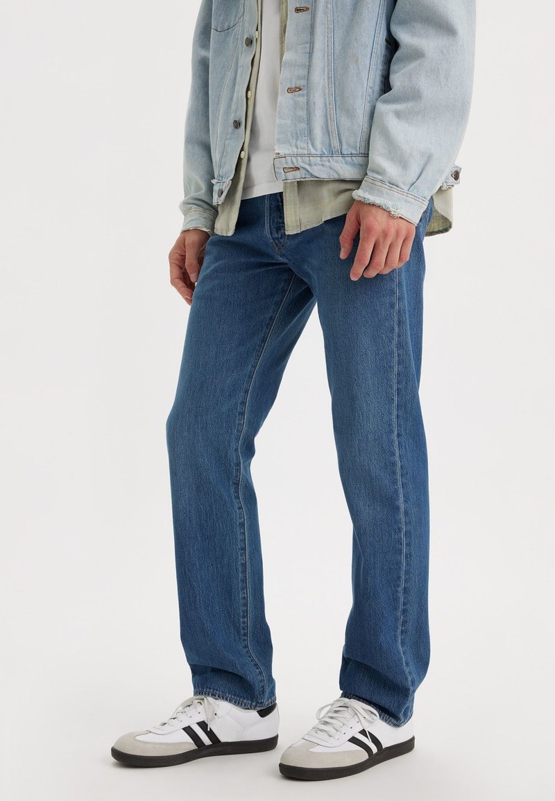 Hellblaue Jeans mit geradem Schnitt, ausgestattet mit fünf Taschen und dezenten Stickdetails, kombiniert mit weißen und schwarzen Sportschuhen.