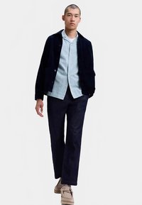 Chaqueta de pana azul marino sobre una camisa azul claro con estampado, combinada con pantalones oscuros y zapatos beige, presentando un look moderno y en capas.