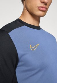 Nike treningsskjorte med blå kropp og svarte skuldre, glatt tekstur, og med en fremtredende oransje swoosh-logo på brystet.