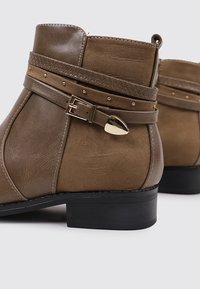 Botines marrones de ante sintético con una correa texturizada y un hebilla en tono dorado. Tacón corto y tachuelas decorativas.