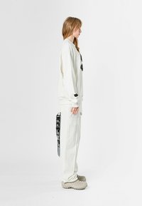 Sudadera blanca con gráfico negro en el frente, combinada con pantalones de chándal a juego que tienen bolsillos laterales y etiquetas. Calzado beige texturizado.