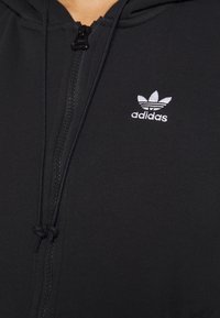 Svart zip-up hoodie med en texturerad tyg, med en liten vit Adidas-logga på bröstet och justerbara snoddar på huvan.