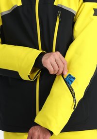 Gelbe und schwarze wasserdichte Jacke mit reflektierenden Akzenten. Verfügt über eine mit Reißverschluss versehenen Armfutter und eine glatte, isolierte Oberfläche. Logo sichtbar.