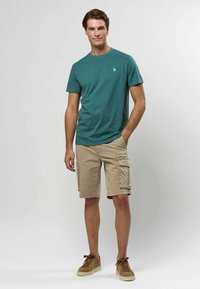 Mėlynai žalias apvalios iškirptės marškinėliai, trumpomis rankovėmis, derinami su chaki spalvos kargo šortais, turinčiais šonines kišenes. Modelis dėvi rudus zomšinius sportbačius.