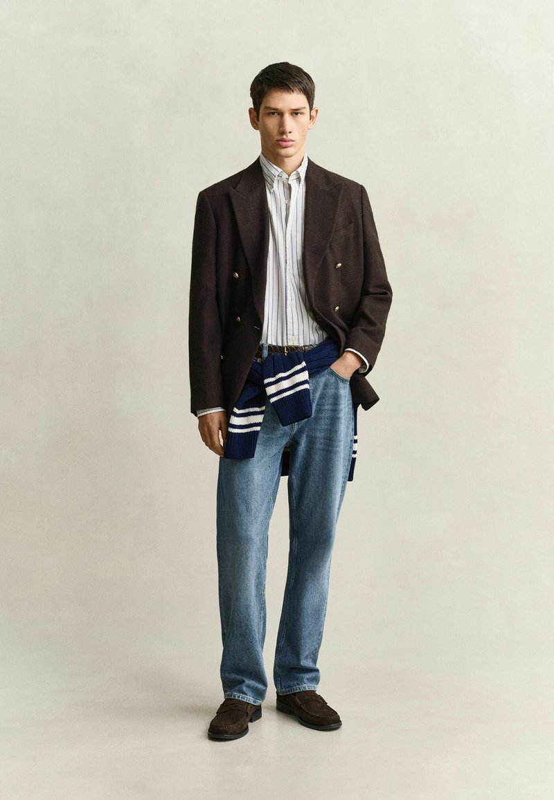Young man stands facing forward, wearing a dark blazer, striped shirt, blue jeans, brown shoes, and a navy sweater tied at his waist.