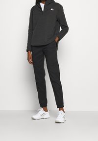 Pullover grigio scuro con mezza zip e logo, abbinato a pantaloni neri a sigaretta. Scarpe sportive bianche con dettagli grigi. Design minimalista.