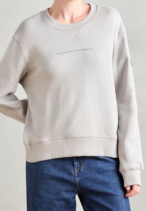 Person iført en beige sweatshirt med teksten "fordi der ikke er nogen planet B" og blå jeans, stående med den ene hånd på hoften.