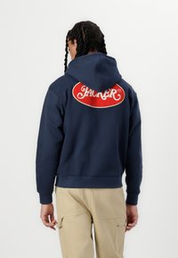 Marinblå hoodie med dragsko i huvan och broderad röd logotyp på baksidan. Mjuk tyg med ribbade ärmslut och nederkant, kombinerad med beige cargobyxor.