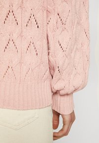 Sweter dziergany w diamentowy wzór w delikatnym różu, z prążkowanym wykończeniem i bufiastymi rękawami, zestawiony z jasnymi spodniami.