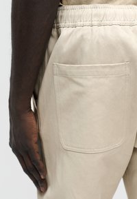 Main à la peau foncée reposant à côté d'une poche à taille élastique beige sur un pantalon de couleur claire, sur un fond neutre.