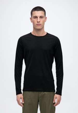 Un homme se tient debout face à l’avant sur un fond clair uni. Il porte une chemise noire à manches longues et un pantalon olive.