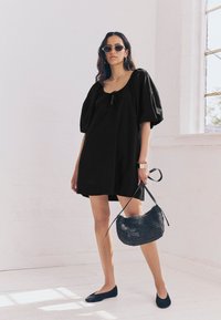 Sort kjole med puffærmer, firkantet udskæring og snøredetalje, kombineret med sorte balletsko og en tekstureret sort flettet håndtaske.