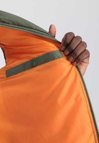 Grünes Jackeninnere mit leuchtend orangefarbener Fütterung, ausgestattet mit einem Reißverschluss und einem kleinen grünen Stoffanhänger mit einem Logo über einer Tasche.