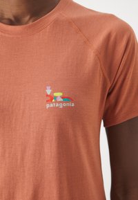 Camiseta de manga corta y tejido ligero en color óxido, con un logotipo colorido de Patagonia en el área izquierda del pecho. Textura de tela suave, cuello redondo.