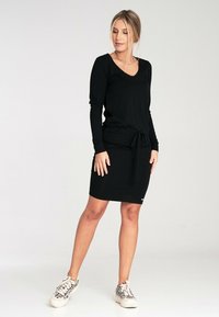 Czarna sukienka z długimi rękawami, dekoltem w serek, cinchowanym w pasie i prostym kroju, zestawiona z wzorzystymi trampkami. Gładka faktura materiału.