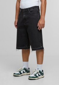 Svart denimshorts med frynset kant, rett passform, fem-lomme design og en lett falming. Kombinert med hvite ankelstrømper og grønne joggesko.