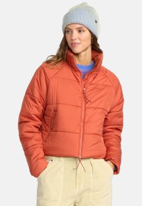 Warme, donzige oranje jas met ritssluiting en zakken, gecombineerd met een lichtblauwe trui en een gestreepte gebreide beanie. Getextureerde stof.