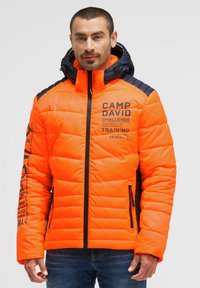Man wearing bright orange and navy padded jacket with hood, printed text on chest and sleeve, and zipped pockets, standing against white background.