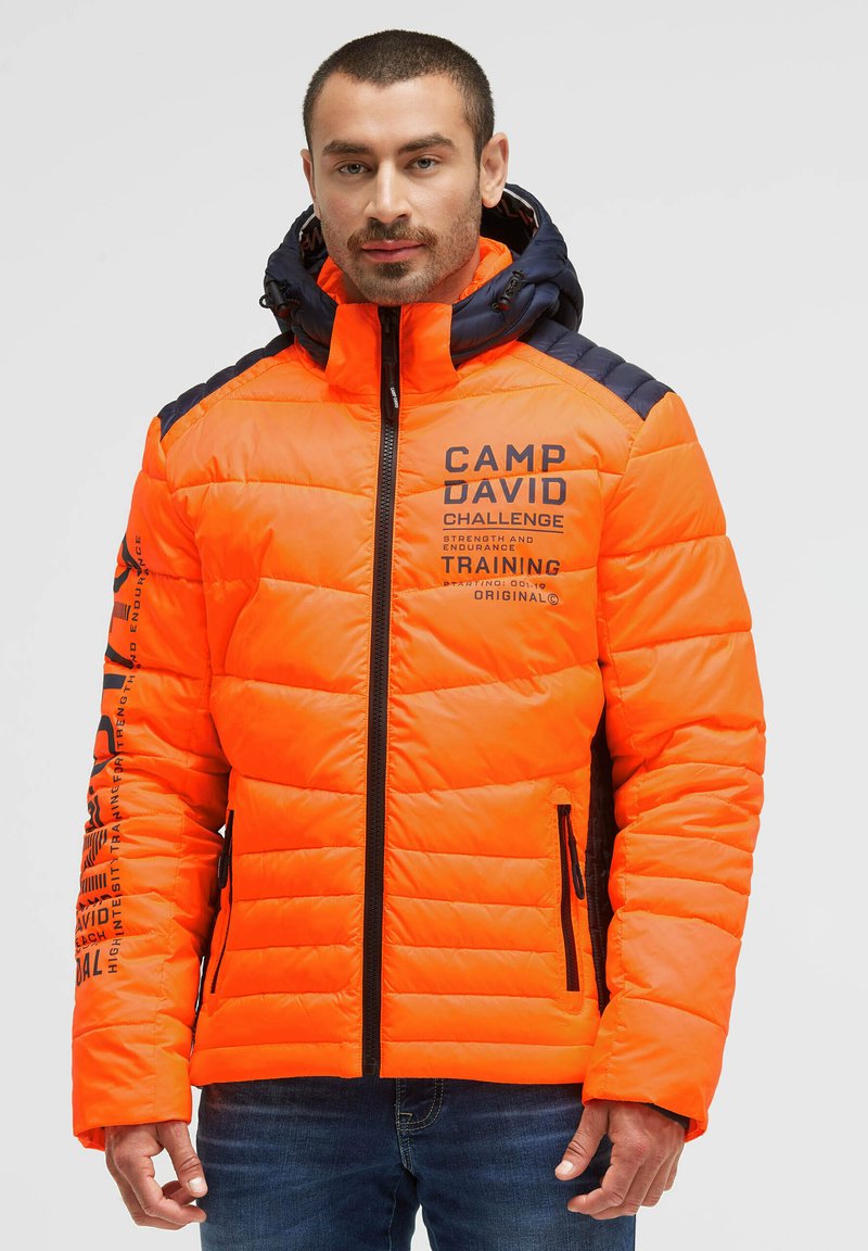 Man wearing bright orange and navy padded jacket with hood, printed text on chest and sleeve, and zipped pockets, standing against white background.
