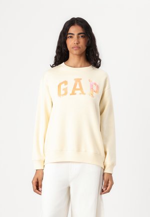 Cream sweatshirt featuring a large, glossy "GAP" logo in orange and pink. Relaxed fit with ribbed cuffs and hem. Worn with white pants.
