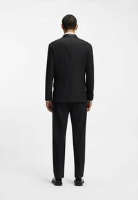 Completo sartoriale nero con giacca aderente e pantaloni dritti. Tessuto liscio, spacco singolo e scarpe eleganti nere. Senza motivi visibili.