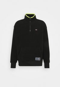 Svart fleece-pullover med halvdragkedja, kontrasterande neongrön innerkrage, framficka och logopatch på nedre vänstra sidan.