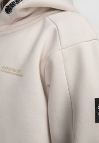 Beige capuchon sweatshirt van zacht materiaal, met een textuur, een geborduurd logo in goud en een zwart accent op de mouw.