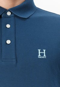 Ναυτικό μπλε πόλο μπλούζα με υφή, διαθέτοντας κλασικό γιακά και τρία κουμπιά. Στο αριστερό στήθος παρουσιάζεται ένα κεντημένο λογότυπο «H» σε ανοιχτό μπλε χρώμα.
