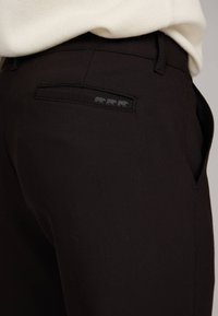 Pantalon noir avec une texture lisse, présentant une poche arrière avec un logo à trois chiffres discret. Design élégant avec une coupe ajustée.