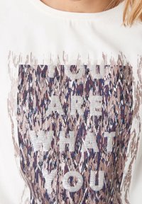 Bílá košile s abstraktním hnědým a modrým vzorem a textem z rhinestone "Jste to, co" na přední straně.