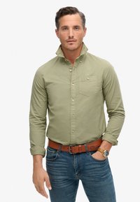 Camisa de botones color verde oliva con mangas largas y un bolsillo en el pecho, combinada con jeans azules y un cinturón marrón. Tejido suave, corte casual.