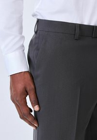 Pantaloni gri închis cu o țesătură texturată, având un croi drept, curea și un buzunar lateral. Materialul pare neted și profesional.