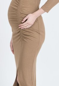 Dopasowana, długa brązowa sukienka z marszczeniem po bokach; charakteryzuje się wysokim rozcięciem i miękkim, elastycznym materiałem. Gładka tekstura, długie rękawy.