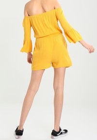 Gul off-shoulder jumpsuit med trumpetärmar, elastisk midja och korta ben. Tyget har en strukturerad yta. Matchas med svarta sneakers.