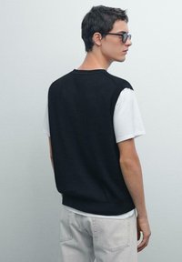 Maglione senza maniche nero lavorato a maglia sopra una maglietta bianca, abbinato a pantaloni grigio chiaro, visto da dietro. Design minimalista e dettagli di texture.