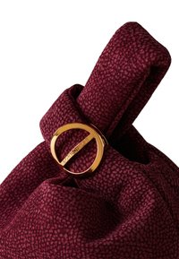 Burgunderfarbene Stofftasche mit strukturierter Oberfläche, versehen mit einem goldenen, runden Hardware-Element. Die Tasche hat ein weiches, gerafftes Oberteil.