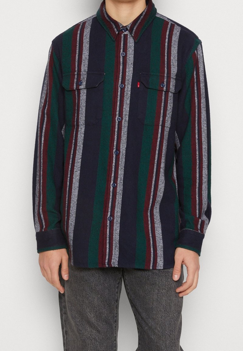 Homme portant une chemise à manches longues boutonnée avec des rayures verticales bleu marine, vert, bordeaux et gris, associée à un jean gris foncé.