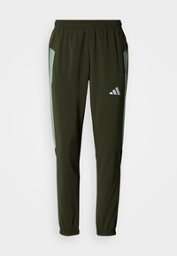 Calças de treino cor verde-oliva com painéis laterais em verde claro, cintura elástica, dois bolsos laterais e um logótipo refletor na coxa esquerda.