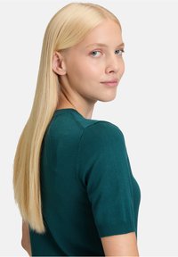 Teal gebreide trui met korte mouwen en een getailleerd ontwerp, geribbelde manchetten en gedetailleerde schoudernaden. Het haar is lang en steil.