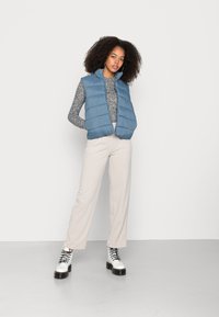 Colete puffer azul claro com fecho de zíper, combinado com uma blusa de manga longa cinza com padrão e calças largas creme. Botas de plataforma pretas com atacadores.